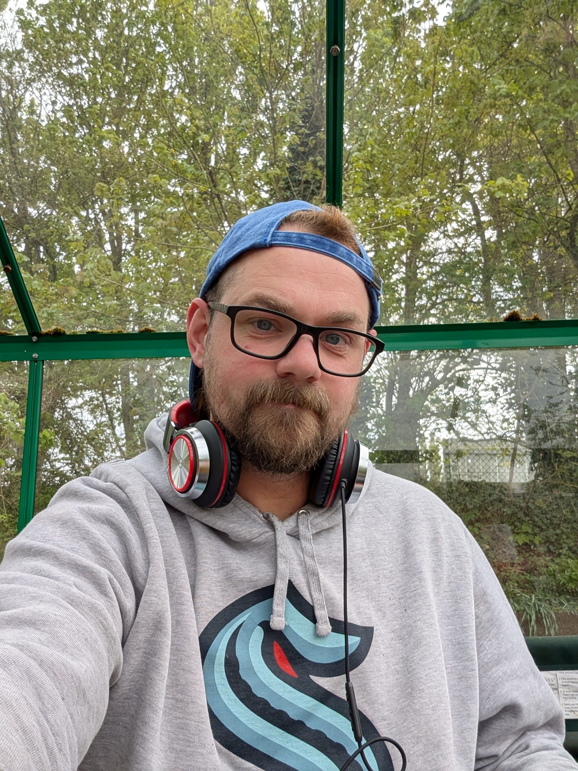 Mike, a chubby white person with a beard is sat inside a greenhouse, wearing a blue baseball cap backwards, square, black rimmed glasses, headphones around their neck, and a grey Seattle Kraken hoodie.